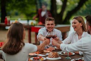 Group of happy friends toasting red wine glass while having picnic french dinner party outdoor during summer holiday vacation near the river at beautiful nature photo