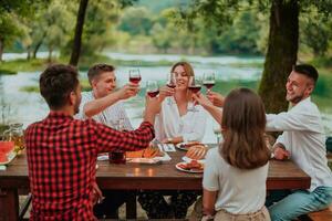 Group of happy friends toasting red wine glass while having picnic french dinner party outdoor during summer holiday vacation near the river at beautiful nature photo