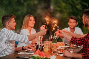 grupo de contento amigos celebrando fiesta vacaciones utilizando aspersores y Bebiendo rojo vino mientras teniendo picnic francés cena fiesta al aire libre cerca el río en hermosa verano noche en naturaleza foto