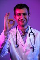 Doctor doing ok sign with fingers, excellent symbol neon lights blue and pink background. Coronavirus pandemic photo