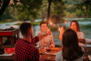 grupo de contento amigos celebrando fiesta vacaciones utilizando aspersores y Bebiendo rojo vino mientras teniendo picnic francés cena fiesta al aire libre cerca el río en hermosa verano noche en naturaleza foto