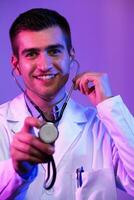 Portrait of hero in white coat. Cheerful smiling young doctor with stethoscope in medical hospital standing against blue and pink background. Coronavirus covid-19 danger alert photo