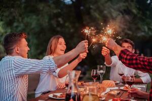 grupo de contento amigos celebrando fiesta vacaciones utilizando aspersores y Bebiendo rojo vino mientras teniendo picnic francés cena fiesta al aire libre cerca el río en hermosa verano noche en naturaleza foto