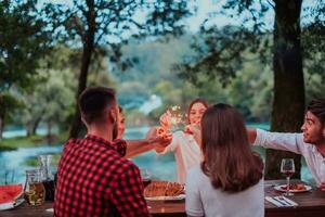 Group of happy friends celebrating holiday vacation using sprinklers and drinking red wine while having picnic french dinner party outdoor near the river on beautiful summer evening in nature photo