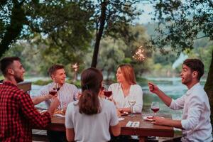 Group of happy friends celebrating holiday vacation using sprinklers and drinking red wine while having picnic french dinner party outdoor near the river on beautiful summer evening in nature photo