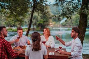 Group of happy friends celebrating holiday vacation using sprinklers and drinking red wine while having picnic french dinner party outdoor near the river on beautiful summer evening in nature photo