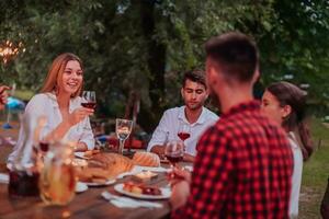 Group of happy friends celebrating holiday vacation using sprinklers and drinking red wine while having picnic french dinner party outdoor near the river on beautiful summer evening in nature photo