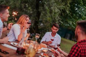 Group of happy friends celebrating holiday vacation using sprinklers and drinking red wine while having picnic french dinner party outdoor near the river on beautiful summer evening in nature photo