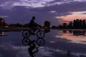 solitario niños silueta en bicicleta, chico montando bicicleta en reflexivo agua. antecedentes hermosa puesta de sol. foto