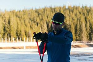 Handsome male athlete with cross country skis, taking fresh breath and having break after hard workout training in a snowy forest. Checking smartwatch. Healthy winter lifestyle. photo