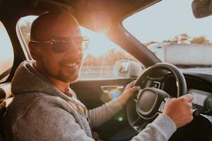 un hombre con un Gafas de sol conducción un coche a puesta de sol. el concepto de coche viaje foto