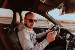 A man with a sunglasses driving a car and type a message on smartphone at sunset. The concept of car travel photo