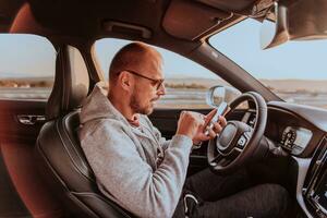 A man with a sunglasses driving a car and type a message on smartphone at sunset. The concept of car travel photo