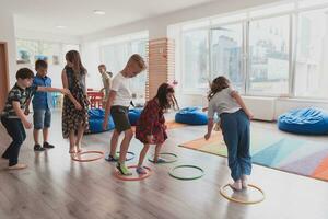 pequeño guardería colegio niños con hembra profesor en piso adentro en aula, haciendo ejercicio. saltando terminado hula aro círculos pista en el piso. foto