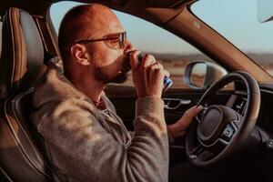 A tired man drinking acoffee while driving a car at sunset. Tired travel and long drive photo