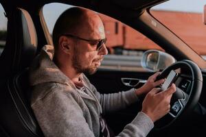 A man with a sunglasses driving a car and type a message on smartphone at sunset. The concept of car travel photo