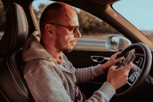 un hombre con un Gafas de sol conducción un coche y tipo un mensaje en teléfono inteligente a puesta de sol. el concepto de coche viaje foto