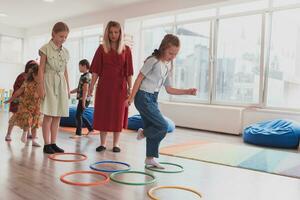 pequeño guardería colegio niños con hembra profesor en piso adentro en aula, haciendo ejercicio. saltando terminado hula aro círculos pista en el piso. foto