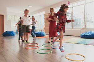 pequeño guardería colegio niños con hembra profesor en piso adentro en aula, haciendo ejercicio. saltando terminado hula aro círculos pista en el piso. foto