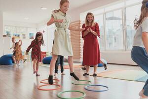 pequeño guardería colegio niños con hembra profesor en piso adentro en aula, haciendo ejercicio. saltando terminado hula aro círculos pista en el piso. foto