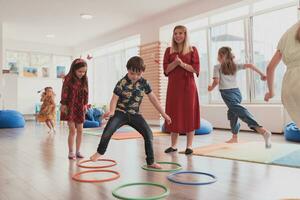 pequeño guardería colegio niños con hembra profesor en piso adentro en aula, haciendo ejercicio. saltando terminado hula aro círculos pista en el piso. foto