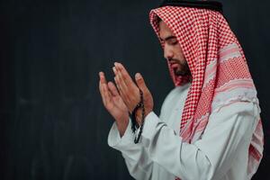 Arabian man in traditional clothes making traditional prayer to God, keeps hands in praying gesture in front of black chalkboard representing modern islam fashion and ramadan kareem concept photo