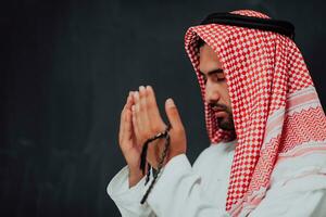 Arabian man in traditional clothes making traditional prayer to God, keeps hands in praying gesture in front of black chalkboard representing modern islam fashion and ramadan kareem concept photo