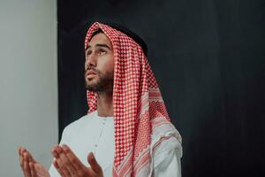 Arabian man in traditional clothes making traditional prayer to God, keeps hands in praying gesture in front of black chalkboard representing modern islam fashion and ramadan kareem concept photo