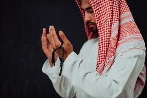Arabian man in traditional clothes making traditional prayer to God, keeps hands in praying gesture in front of black chalkboard representing modern islam fashion and ramadan kareem concept photo