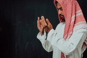 Arabian man in traditional clothes making traditional prayer to God, keeps hands in praying gesture in front of black chalkboard representing modern islam fashion and ramadan kareem concept photo