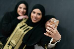 Group of young beautiful muslim women in fashionable dress with hijab using smartphone while taking selfie picture in front of black background photo