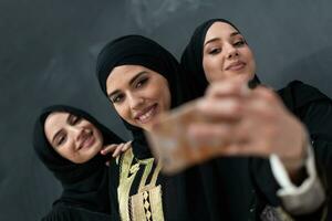 grupo de joven hermosa musulmán mujer en de moda vestir con hijab utilizando teléfono inteligente mientras tomando selfie imagen en frente de negro antecedentes foto