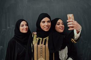 grupo de joven hermosa musulmán mujer en de moda vestir con hijab utilizando teléfono inteligente mientras tomando selfie imagen en frente de negro antecedentes foto