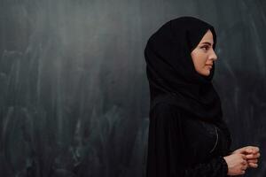 Portrait of beautiful muslim woman in fashionable dress with hijab in front of black chalkboard representing modern islam fashion and ramadan kareem concept photo