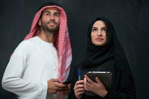 Young muslim business couple arabian man with woman in fashionable hijab dress using mobile phone and tablet computer in front of black chalkboard representing modern islam fashion technology photo
