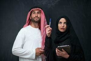 Young muslim business couple arabian man with woman in fashionable hijab dress using mobile phone and tablet computer in front of black chalkboard representing modern islam fashion technology photo