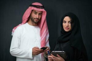 Young muslim business couple arabian man with woman in fashionable hijab dress using mobile phone and tablet computer in front of black chalkboard representing modern islam fashion technology photo