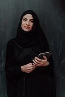 joven moderno musulmán negocio mujer utilizando tableta computadora vistiendo lentes y hijab ropa en frente de negro pizarra foto