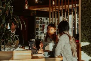 two young business women sitting at table in cafe. Girl shows colleague information on laptop screen. Girl using smartphone, blogging. Teamwork, business meeting.. photo