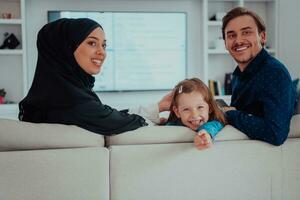 contento musulmán familia con hija mujer en tradicional de moda vestir teniendo divertido y bueno hora juntos mientras sentado en sofá foto