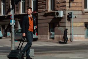 Going to airport terminal. Confident businessman traveler walking on city streets and pulling his suitcase drinking coffee and speaking on smartphone photo