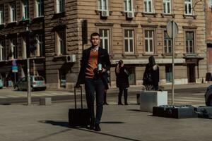 ir a la terminal del aeropuerto. viajero de negocios confiado caminando por las calles de la ciudad y tirando de su maleta bebiendo café y hablando en el teléfono inteligente foto