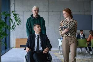 A group of business colleagues in a modern office exemplifies inclusivity and support as they compassionately wheel their business friend in a wheelchair, showcasing teamwork and empowerment in the workplace. photo