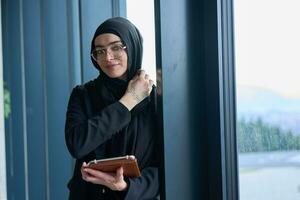 A hijab businesswoman stands by the window in a modern office holding a tablet in her hand showcasing her professionalism, technological prowess and entrepreneurial spiri photo