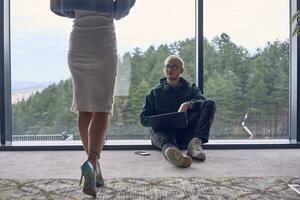 A young blond man in a modern office is sitting by the window, engrossed in his work on a laptop while talking to a female colleague photo