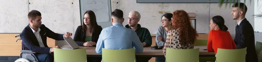 Wide crop photo of a diverse group of business professionals, including an person with a disability, gathered at a modern office for a productive and inclusive meeting.