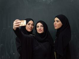 grupo de joven hermosa musulmán mujer en de moda vestir con hijab utilizando teléfono inteligente mientras tomando selfie imagen en frente de negro antecedentes foto