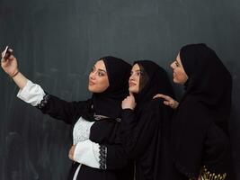 Group of young beautiful muslim women in fashionable dress with hijab using smartphone while taking selfie picture in front of black background photo