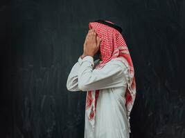 Arabian man in traditional clothes making traditional prayer to God, keeps hands in praying gesture in front of black chalkboard representing modern islam fashion and ramadan kareem concept photo