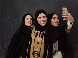 grupo de joven hermosa musulmán mujer en de moda vestir con hijab utilizando teléfono inteligente mientras tomando selfie imagen en frente de negro antecedentes foto